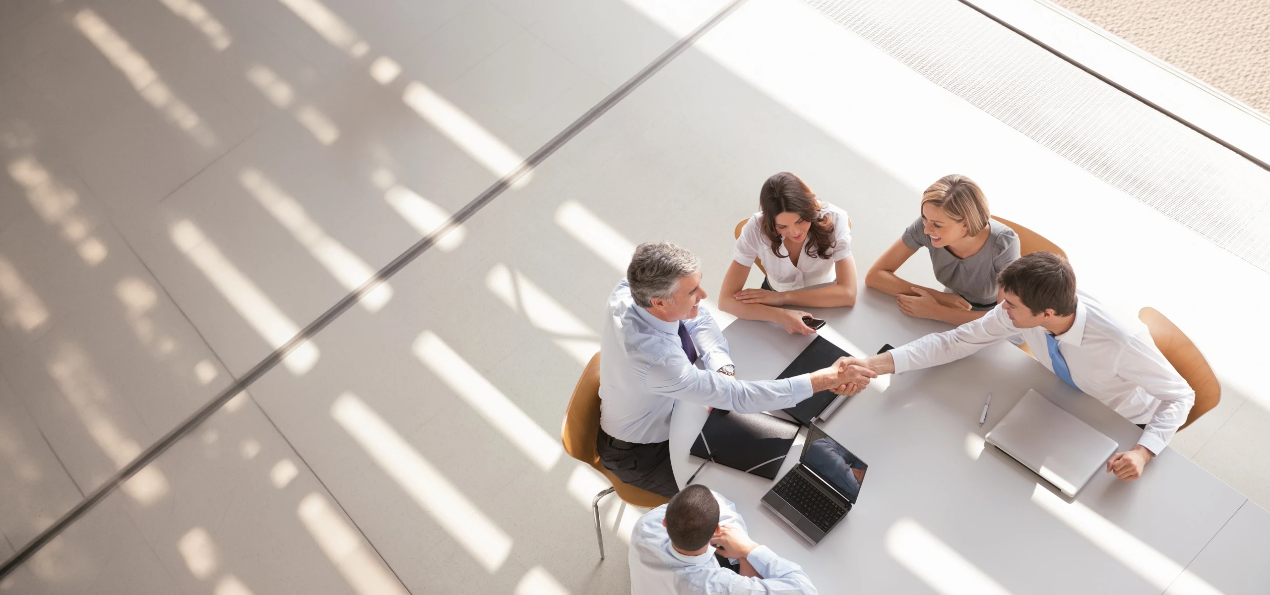 Overhead view of five people at a business meeting, with two participants shaking hands across a table.