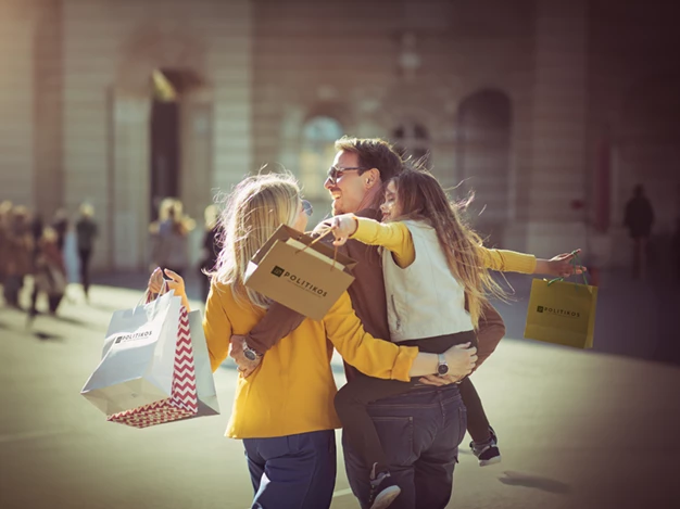 A man is carrying a young girl in his arms, while a woman walks beside them. All three appear to be smiling and in good spirits.They are holding shopping bags, suggesting they’ve been shopping and are enjoying a walk together.