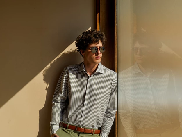 shows a young man standing next to a window or glass surface, where his reflection is visible. He is wearing sunglasses, a striped shirt, and a belt, giving him a neat and stylish appearance.