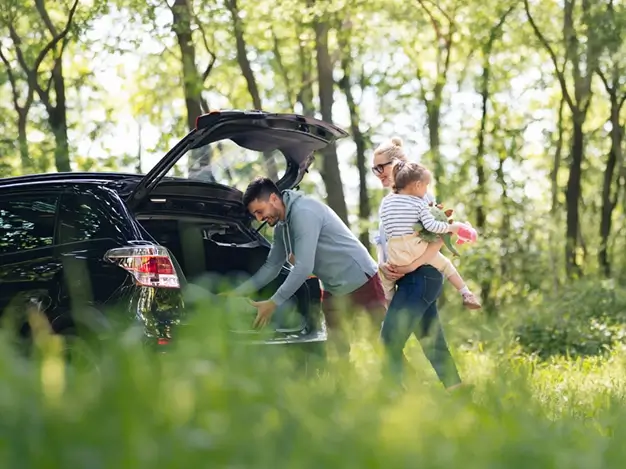 A car is parked among the trees with its trunk open. A man is bending down to take items out of the trunk, while next to him a woman is holding a small child in her arms.