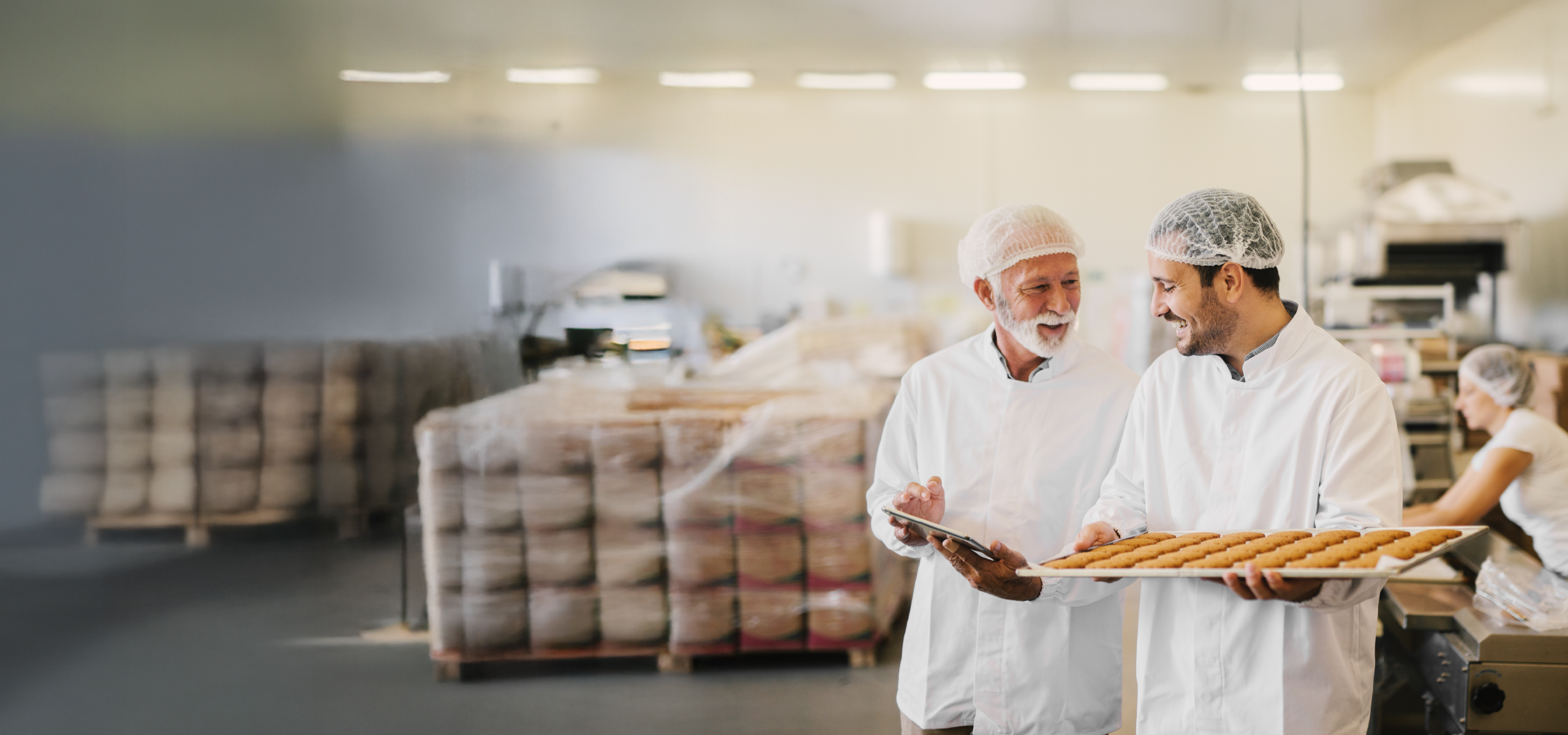 Two people wearing white coats and hairnets hold a tray of baked goods and a tablet while standing in a production or bakery facility; shelves and another worker are visible in the background.