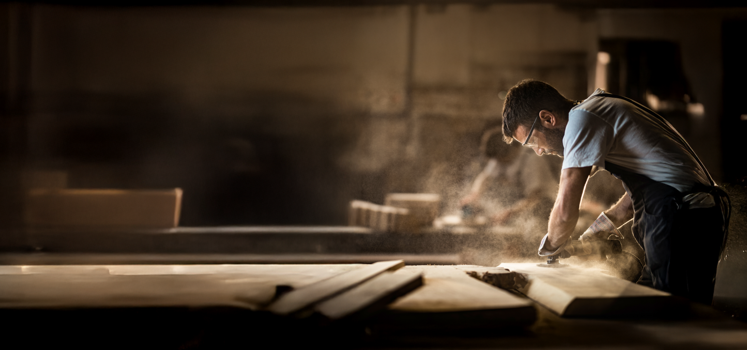 A person wearing safety glasses and gloves sands a wooden surface inside a workshop using a power tool, with fine dust particles illuminated by warm light and machinery blurred in the background.