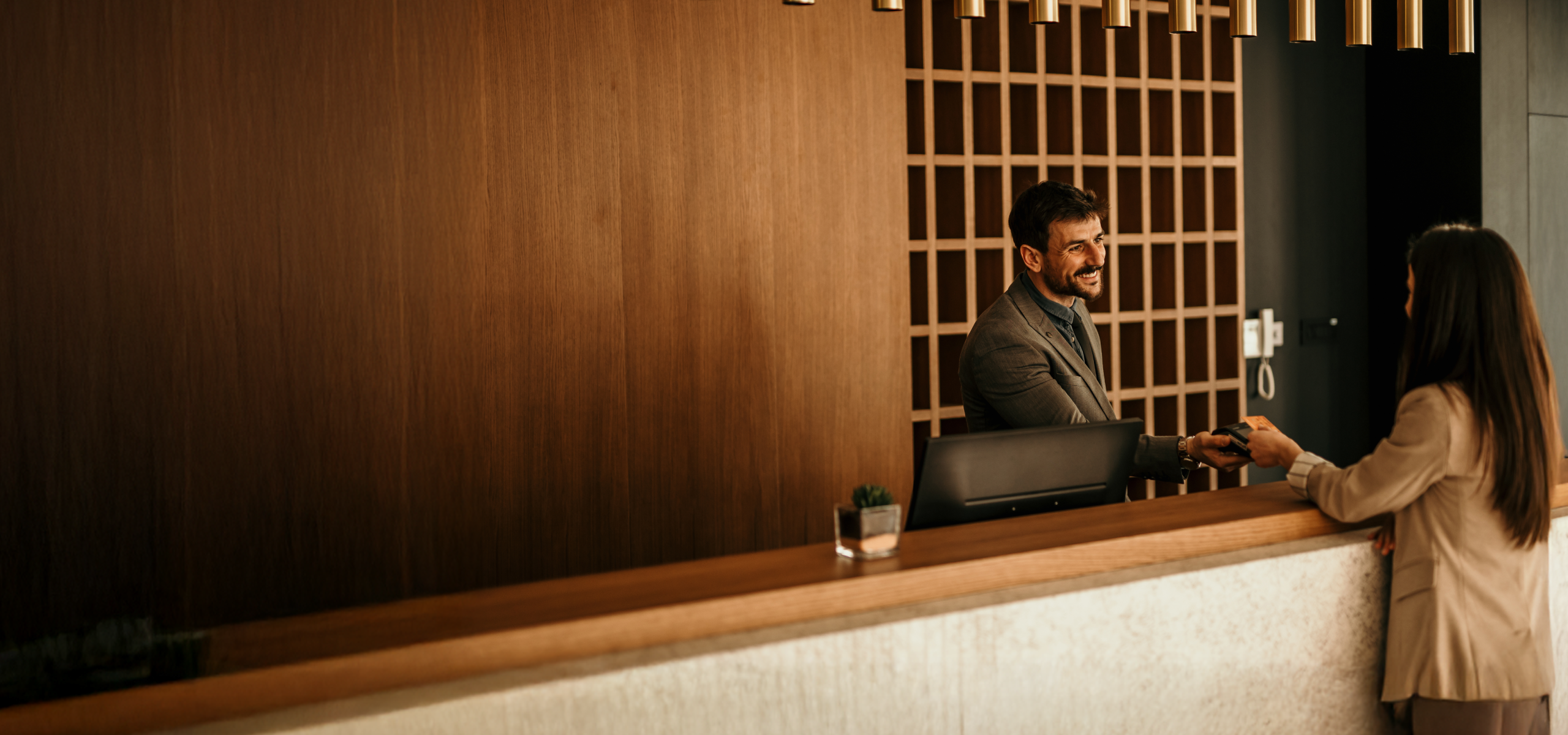 A person standing behind a reception desk hands a card or document to a visitor across the counter in a modern interior space with wood paneling and soft lighting.